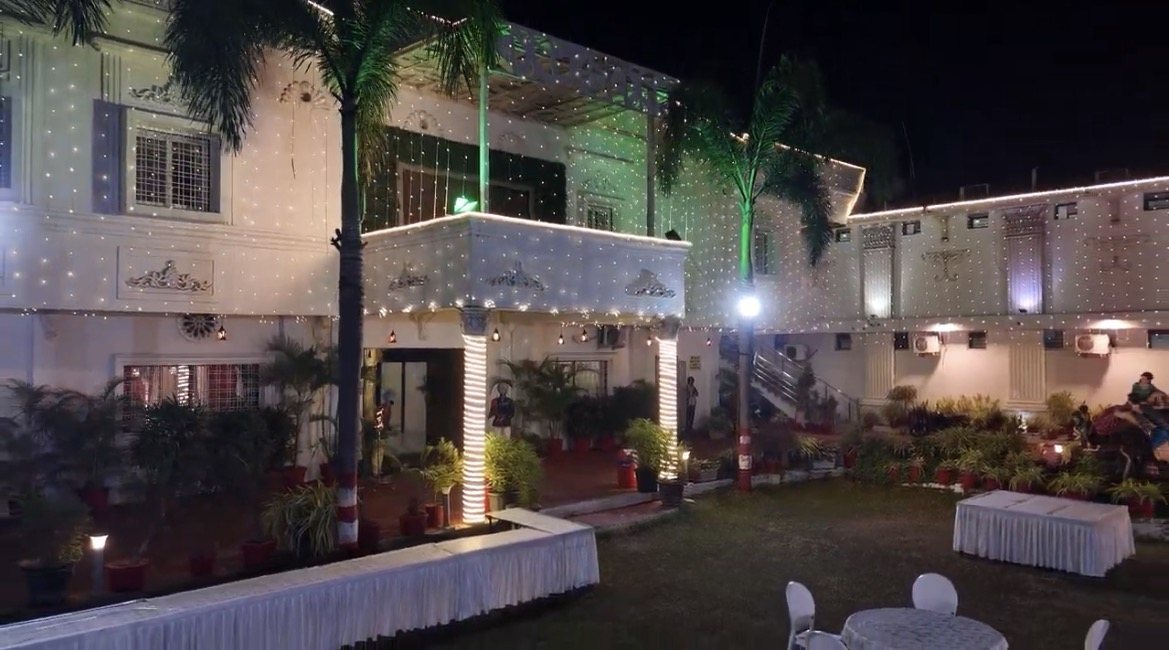 Front of Abhinandan Palace with its illuminated portico, palm trees, garden and lawn seating at night